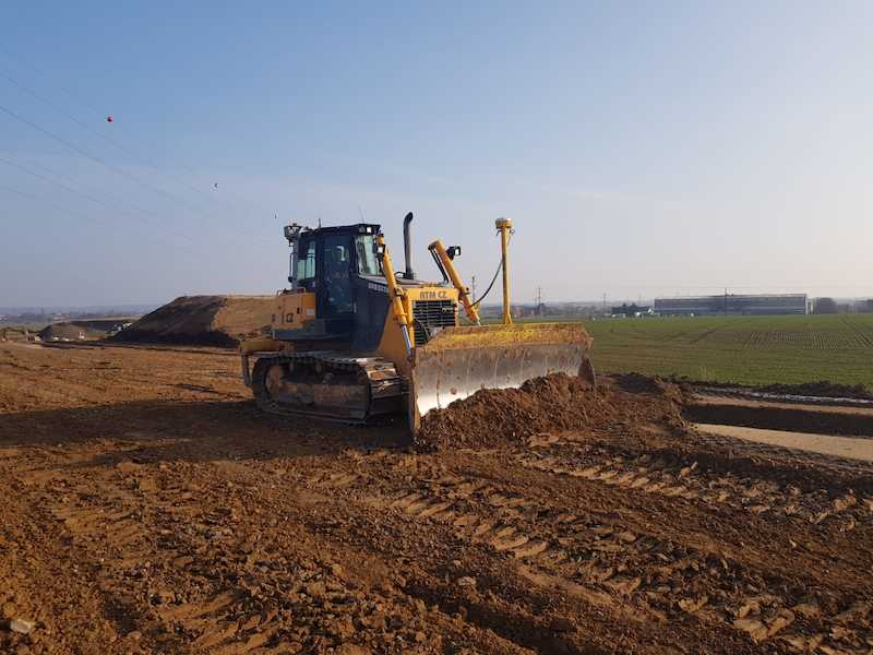 Žlutý buldozer, jeden z nepostradatelných stavebních strojů, odhrnuje zeminu na staveništi pod jasně modrou oblohou, přičemž v pozadí jsou vidět otevřená pole a budova.