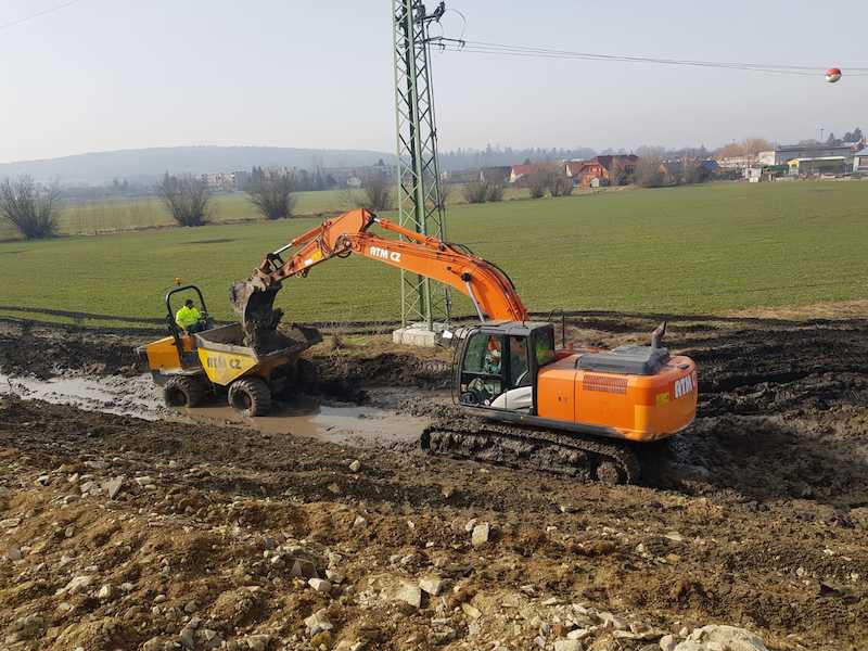 Oranžové rypadlo provádí zemní práce a nakládá zeminu do sklápěče na blátivém staveništi poblíž travnatého pole s stromy, elektrickým vedením a domy v dálce.