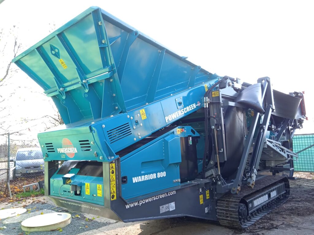 Na staveništi stojí pod širým nebem velký modrý průmyslový třídič s označením POWERSCREEN WARRIOR 800, připravený k zemním pracím a stabilizaci půdy; jsou na něm vidět kovové pásy a různé výstražné štítky.